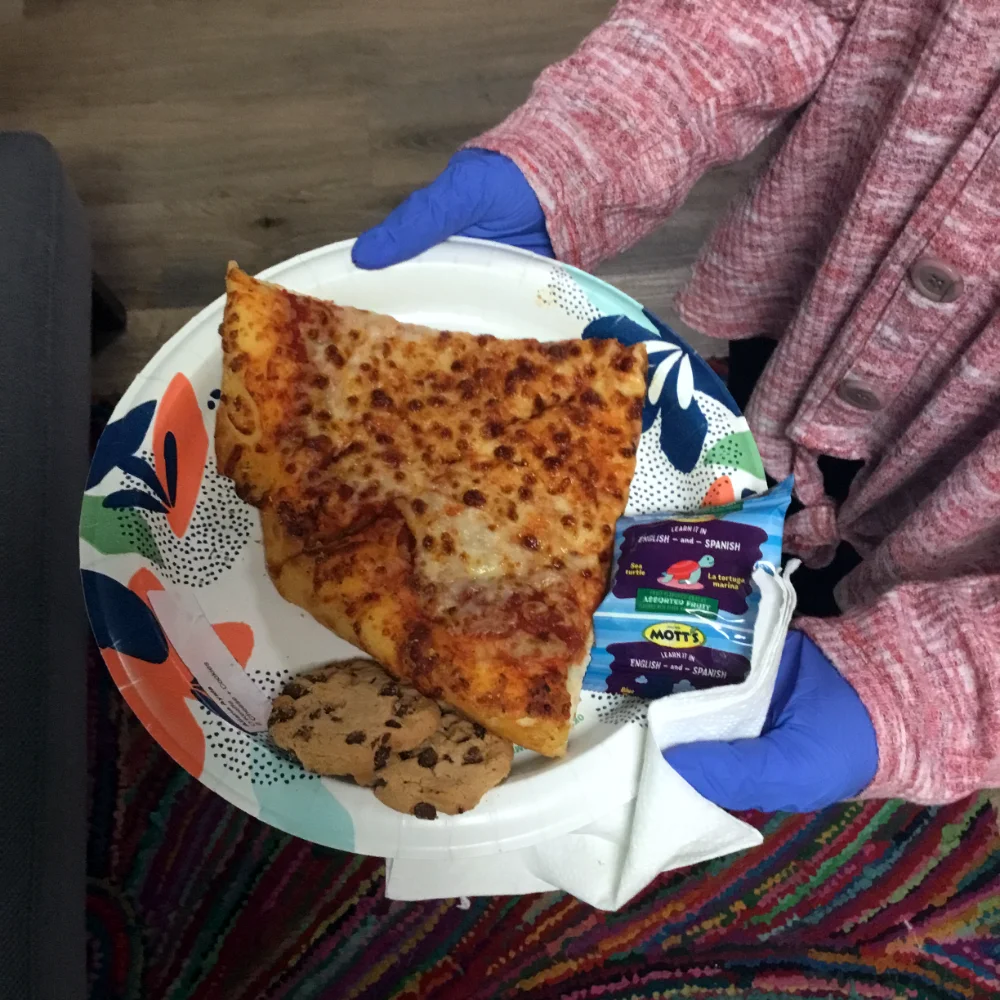 One of the students wearing blue gloves and holding a plate containing two slices of cheese pizza.